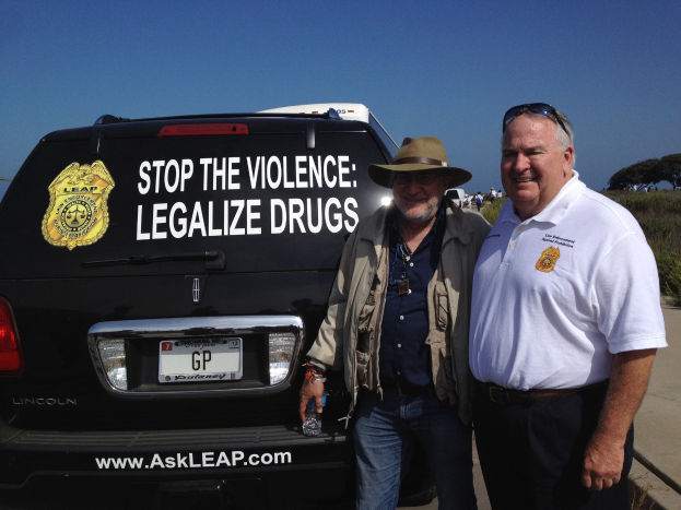 Zwei Männer stehen neben einem schwarzen Van mit "Stop the Violence Legalize Drugs" darauf, einer hölt eine Flasche, mit Menschen, Bäumen und einem klaren blauen Himmel im Hintergrund.
