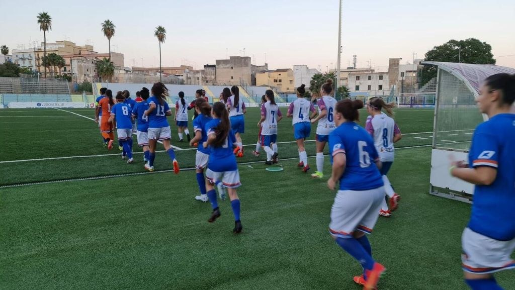 Gruppe junger Frauen beim Fußballspielen auf einem Rasenfeld mit Maschendrahtzaun und einer Wand im Hintergrund, Text in der rechten unteren Ecke lautet "Paris Saint-Germain Frauenfußball".