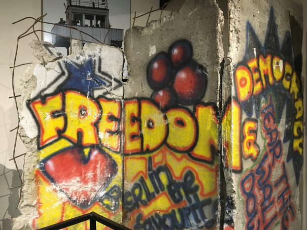 Eine Wand mit k├╝hnem, schwarzem Graffiti, das 'Freiheit und Demokratie' lautet, in einem Raum mit einem Gel├Ąnder unten und einem Fenster im Hintergrund.