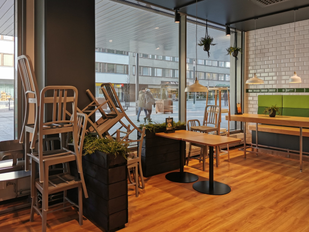 Außensitzbereich eines Berliner Restaurants mit Holztischen und -stühlen, großen Fenstern, Pflanzen und Beleuchtung, mit Blick auf eine Stadtstraße mit Fußgängern, Gebäuden und Strommasten.
