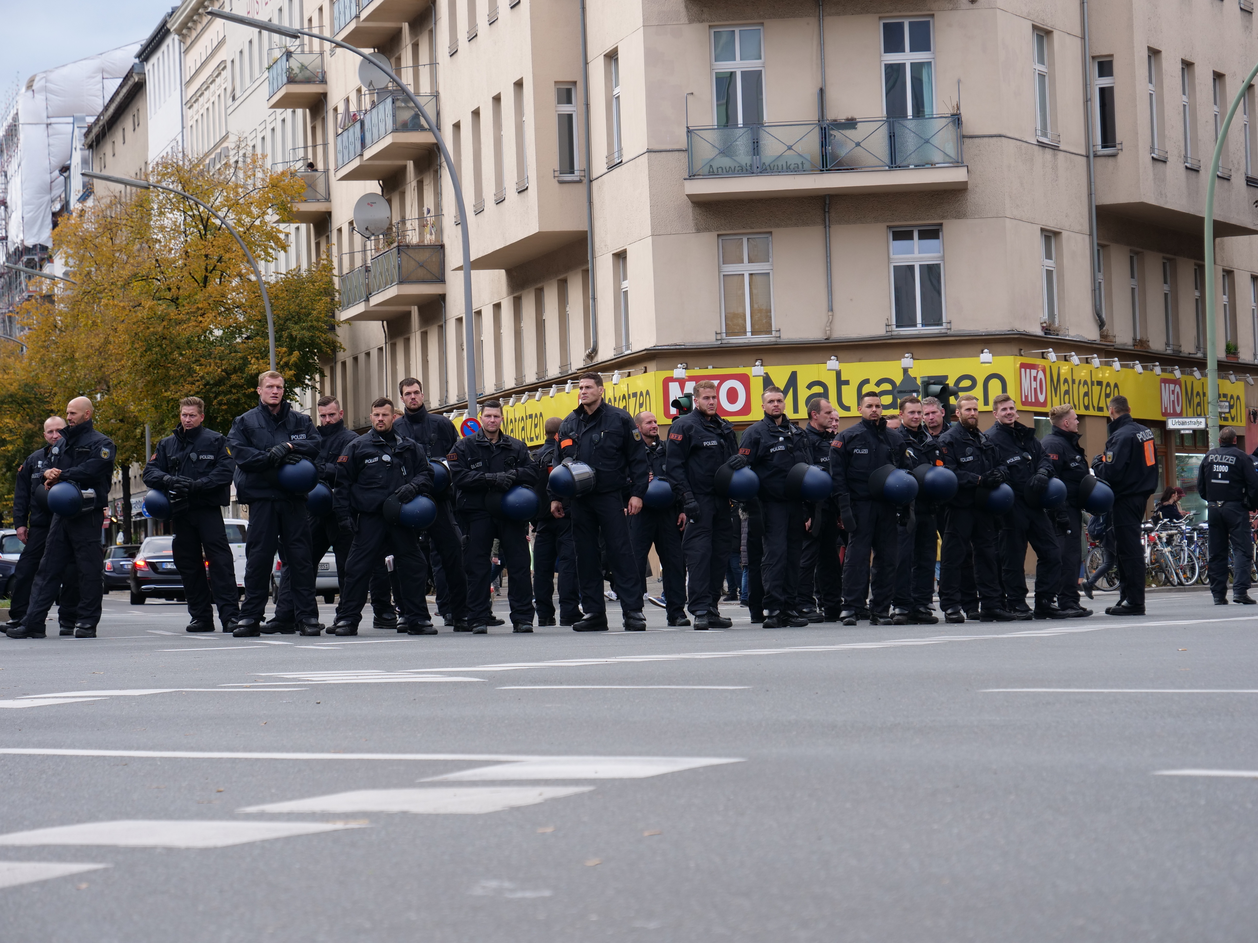 Eine Gruppe von Polizisten in schwarzen Uniformen hält blaue Helme in der Mitte einer Straße, die von Laternenmasten, Bäumen und Gebäuden mit Glasfenstern und Balkonen gesäumt ist, mit weiteren Bäumen und einem klaren blauen Himmel im Hintergrund.