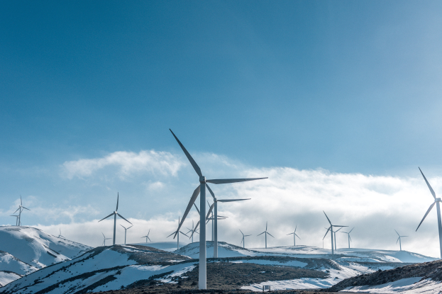 Eine Gruppe von Windkraftanlagen steht in einem verschneiten Feld, mit verschneiten Hügeln und bewölktem Himmel im Hintergrund und Text unten, der ein erneuerbares Energieprojekt angibt.