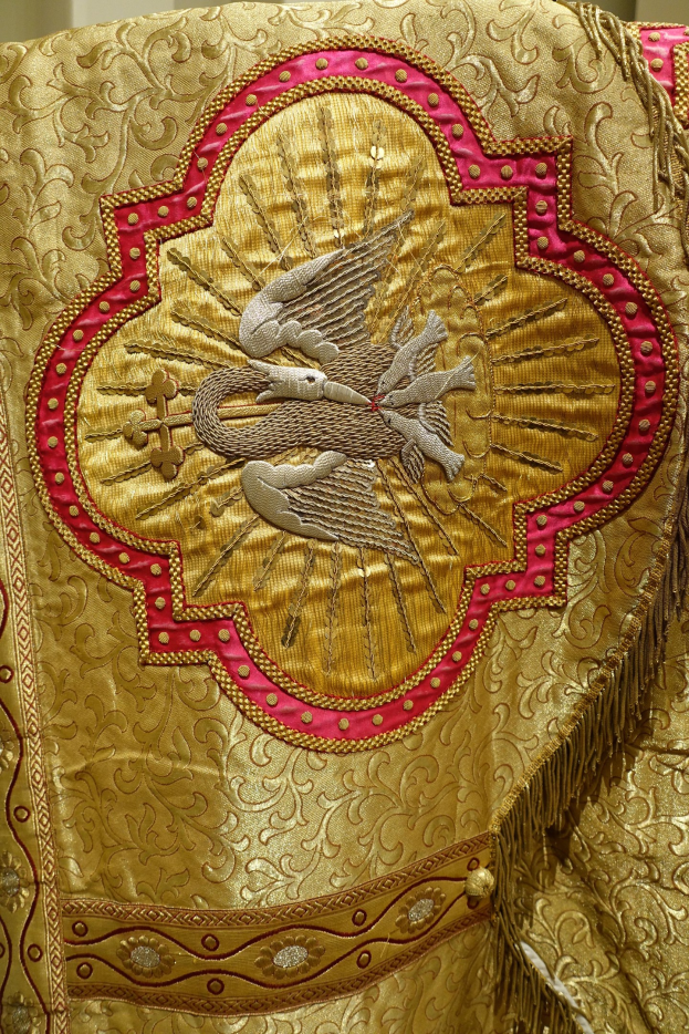 Nahaufnahme eines Priestergewandes mit goldenen und roten Stickereien und detaillierten Mustern, das an einer Wand im Hintergrund hängt.