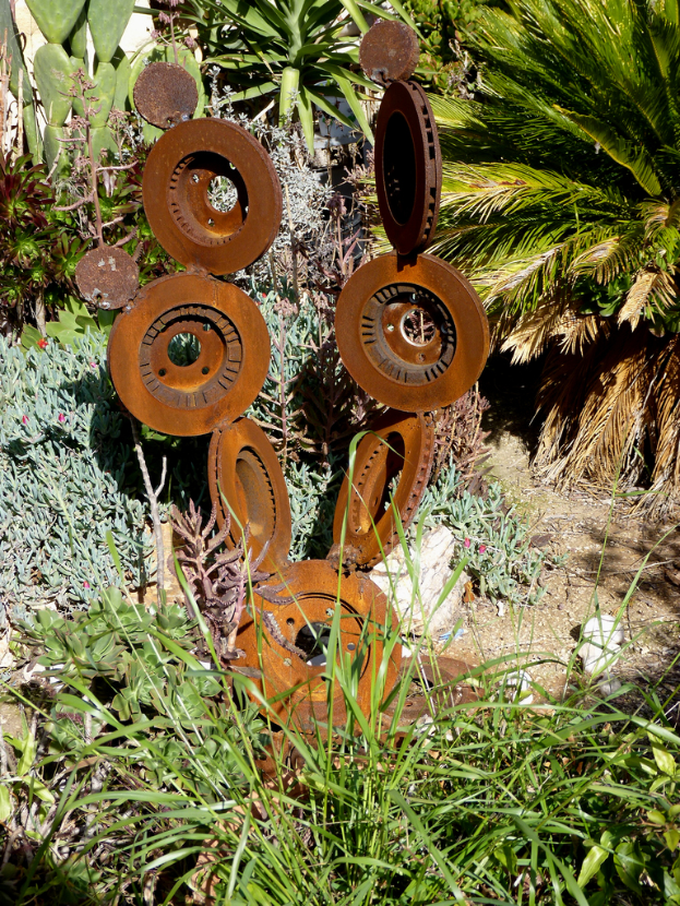 Eine Metallskulptur steht auf einem Stück Land mit spärlicher Vegetation, darunter einige Pflanzen, Grasflecken und verstreute Bäume.