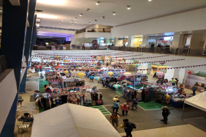 Ein volles Einkaufszentrum mit Kleiderständen, Dekorationsfahnen, Ballons und Menschen, die zwischen Geländern, Säulen und Deckenleuchten sitzen und stehen.