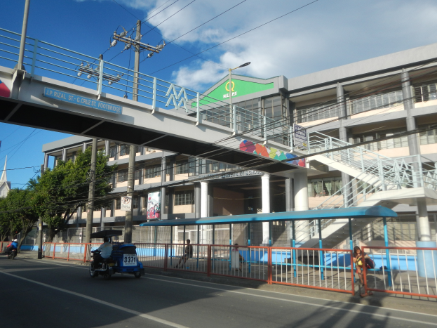 Eine belebte Stadtstraße mit Fahrzeugen, einer Brücke, Strommasten, Laternen, Bäumen, Gebäuden mit Fenstern, Bannern und einem bewölkten Himmel.
