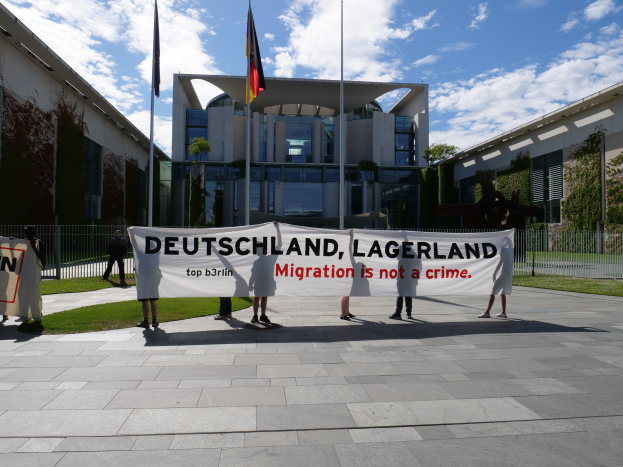 Eine Gruppe von Menschen hält eine Fahne mit der Aufschrift "Deutschland, Lagerland Migration ist kein Verbrechen" vor einem Gebäude mit Fenstern, umgeben von Gras, einem Metallzaun, Pflanzen, Bäumen und Fahnenmasten unter einem bewölkten Himmel.