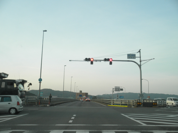 Ein Auto fährt auf einer Autobahn mit einer Ampel an der Kreuzung, umgeben von Fahrzeugen, einem Fußgänger auf dem Gehweg, Geländern, Laternen, Verkehrsschildern, Texttafeln, Gebäuden, Hügeln und einem klaren blauen Himmel.