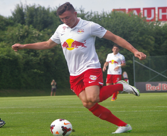 Ein professioneller Fußballspieler schießt einen Ball auf einem grünen Feld mit Bäumen und einem klaren blauen Himmel im Hintergrund, in der Nähe einer Tafel mit der Aufschrift "RB Leipzig."