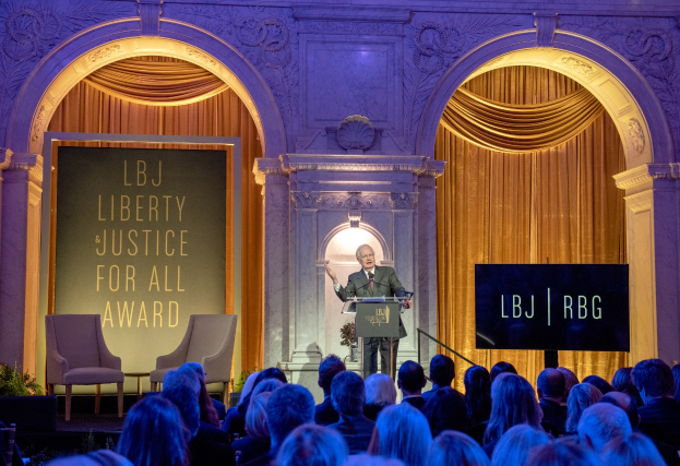 Ein Mann steht an einem Podium mit einem Mikrofon und spricht vor einem Publikum, das auf Stühlen sitzt, in einem Raum mit Pflanzen, Vorhängen und einer Tafel mit Text dahinter.