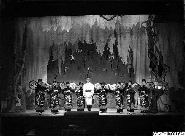 Schwarzes und weißes Bild von Darstellern auf einer Bühne in traditionellen japanischen Kimonos, die Gegenstände halten, mit einer bemalten Kulisse und Text, der unten 'Kimono-Show in Tokyo, Japan' lautet.