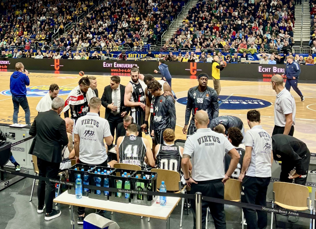 Gruppe von Männern auf einem Basketballfeld während eines Spiels zwischen Virtus Berlin und Echt Berlin, umgeben von sitzenden und stehenden Zuschauern, mit einem Fotografen und Equipment links und Texttafeln im Hintergrund.