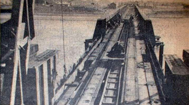Ein Schwarz-Weiß-Foto einer Bahnschiene mit einer im Hintergrund befindlichen Brücke, umgeben von verstreuten Werkzeugen und Materialien.