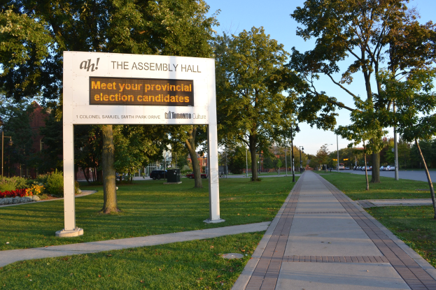 Ein Kreidetafel auf Gras mit der Aufschrift "Die Versammlungshalle - Treffen Sie Ihre Landtagswahlkandidaten" neben einem Weg, Bäumen, Blumen, Straßenlaternen, Fahrzeugen, einem Gebäude und einem bewölkten Himmel.