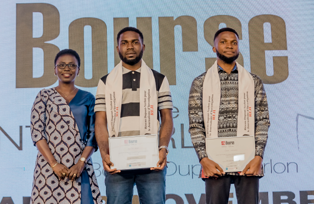 Drei Personen stehen nebeneinander auf einer Bühne, jede hält ein Zertifikat in der Hand, mit einem Bildschirm im Hintergrund, der 'Bounce Africa Awards 2019' anzeigt.
