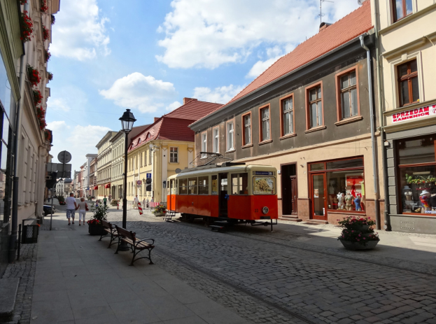 Eine rote und gelbe Straßenbahn fährt durch die Kopfsteinpflasterstraße der Altstadt von Tallinn, flankiert von Gebäuden mit Fenstern, Bänken und Laternen, mit Passanten auf den Gehwegen und einem bewölkten Himmel im Hintergrund.