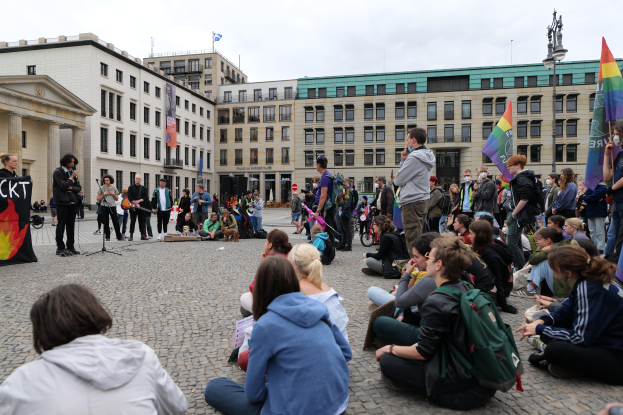 Eine Gruppe von Menschen sitzt auf dem Boden vor einer Menge, die Fahnen und Plakate hält, mit einer Person, die ein Mikrofonständer, ein Plakat, eine Statue auf einem Sockel und Gebäude mit Fenstern im Hintergrund hält, während eine anti-schwulen Demonstration in Berlin, Deutschland, unter einem bewölkten Himmel stattfindet.
