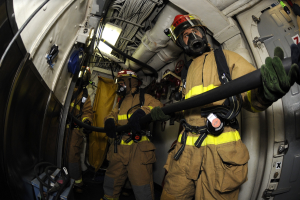 Feuerwehrleute in Helmen und Handschuhen mit einer Schlauchleitung in einem Flugzeug, mit Ausrüstung und Text im Hintergrund.