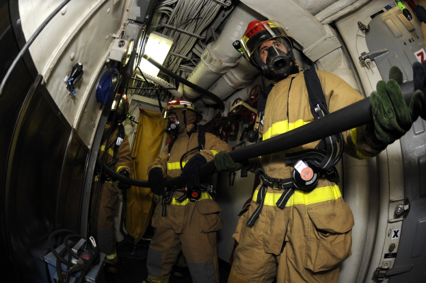 Feuerwehrleute in Helmen und Handschuhen mit einer Schlauchleitung in einem Flugzeug, mit Ausrüstung und Text im Hintergrund.
