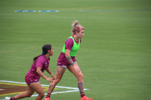 Zwei Frauen beim Fussballspielen auf einem Feld, eine trägt ein grünes T-Shirt und Shorts, die andere ein pinkes Kleid, beide haben Tätowierungen an Händen und Füssen.