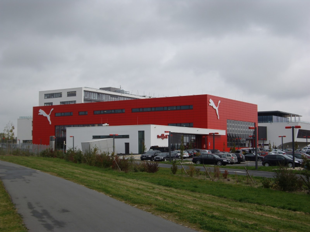 Übersicht des Puma-Werks in Deutschland mit Gebäuden, Fahrzeugen, Straßeninfrastruktur, Vegetation und einem bewölkten Himmel.