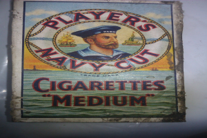 Plakat, das "Player's Navy Cut Cigarettes Medium" bewirbt, zeigt eine Person in einer marineblauen Uniform mit weißem Hemd, roter Krawatte, strenger Miene und verschränkten Armen.