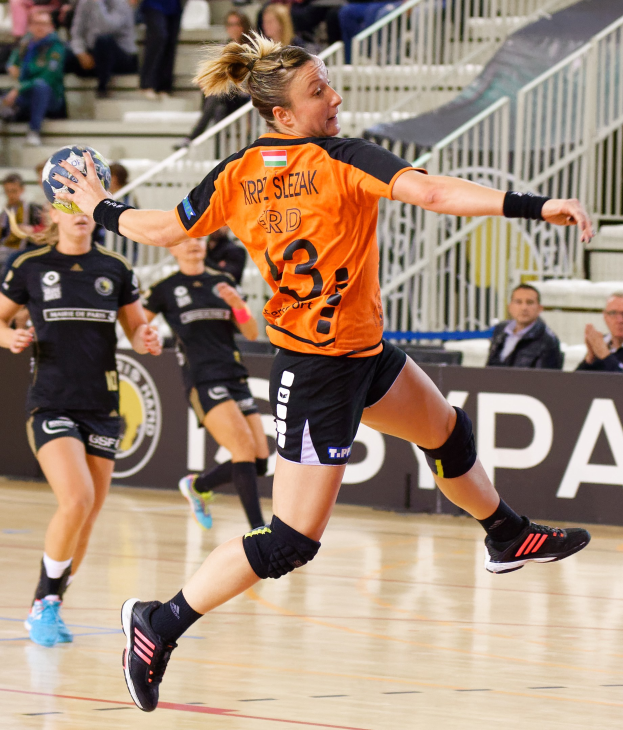 Eine Frau in einem orangen T-Shirt und schwarzen Shorts hält einen Handball, mit Zuschauern auf Treppen und Geländern im Hintergrund während des "Krp Slezak - Handball-Weltmeisterschaft 2019".