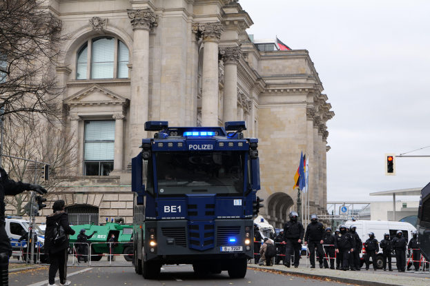 Eine Gruppe von Polizeibeamten steht vor einem grossen Gebäude mit Fenstern, Säulen und Bögen, mit Fahrzeugen auf der Strasse, einer Person mit einer Kamera auf der linken Seite und einem klaren blauen Himmel im Hintergrund.