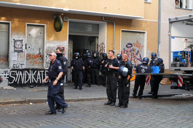 Eine Gruppe von Polizisten, einige mit Helmen und Waffen in der Hand, steht und geht vor einem Graffiti-Gebäude, mit einem Fahrzeug und verschiedenen Gegenständen im Hintergrund.
