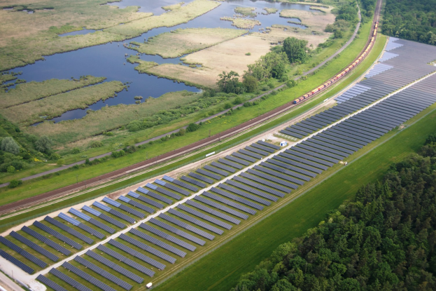 Eine Luftaufnahme eines Solarparks mit zahlreichen Solarpanelen auf einem Feld, umgeben von Bäumen, Gras und Wasser, mit einem Zug, der auf einem nahen Bahngeleis fährt.
