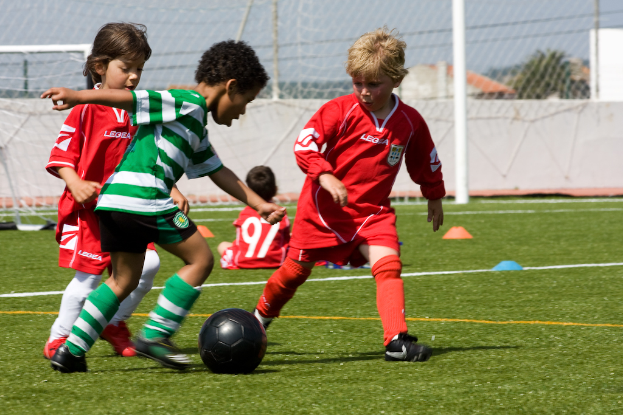 Drei Kinder spielen Fußball auf einem Feld mit einem vierten Kind, das dahinter sitzt und ein Netz im Hintergrund.