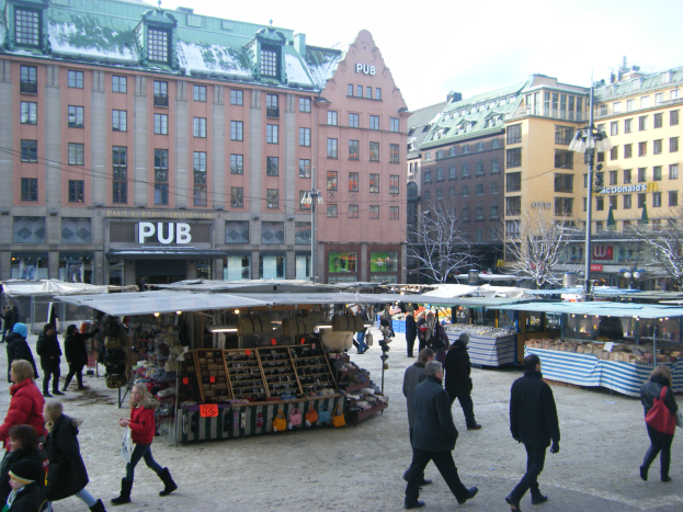 Ein lebendiger Weihnachtsmarkt in Stockholm, Schweden, mit Menschen, die herumlaufen, Ständen voller Gegenstände, Gebäuden, Laternenmasten, Bäumen und einem klaren blauen Himmel im Hintergrund.