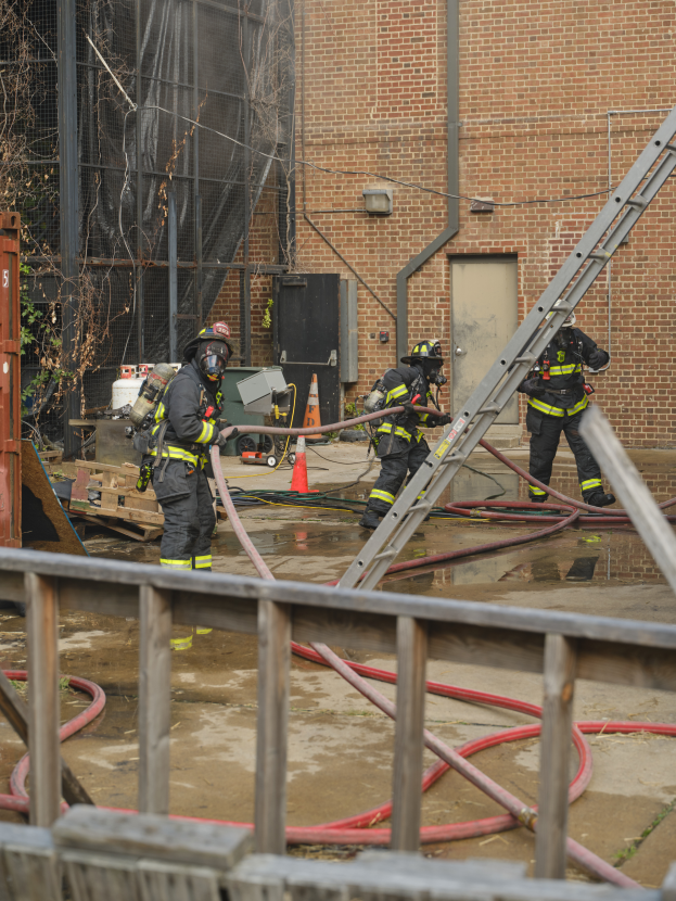 Eine Gruppe von Feuerwehrleuten in Helmen, die Schläuche halten, arbeiten daran, ein Gebäude Feuer zu löschen, mit verschiedenen Ausrüstungen und Strukturen drumherum, einschließlich eines Metallzauns, Schläuchen auf dem Boden, einem Container, einem Verkehrskegel und einem Baum im Hintergrund.