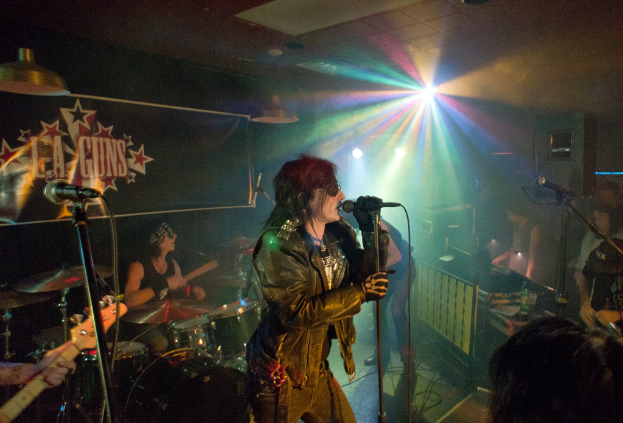 Eine Frau in einer schwarzen Jacke singt auf der Bühne in ein Mikrofon, während ein Mann mit Sonnenbrille und Stirnband ein Instrument spielt und ein Plakat mit der Aufschrift "LA Guns" im Hintergrund zu sehen ist.