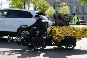 Ein Mann im Rollstuhl mit einem großen Motor auf dem Rücken, umgeben von Fahrzeugen auf einer Straße mit Bäumen, Gebäuden, Polen und einem klaren blauen Himmel im Hintergrund; er trägt eine schwarze Jacke, eine Kappe und hält ein Objekt in der Hand.