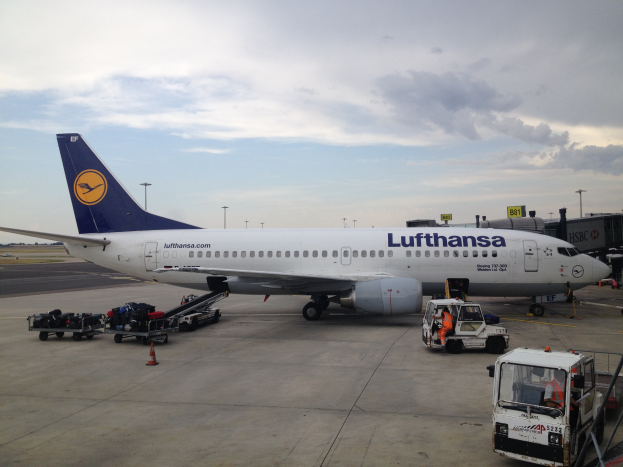 Ein Lufthansa-Airbus A320-200 auf dem Frankfurter Flughafen geparkt, mit verschiedenen Bodenfahrzeugen, Wagen, Verkehrskegeln, Pfählen und Texttafeln im Bild.