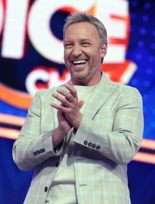 Ein Mann im Anzug, der auf einer Bühne klatscht, mit "The Voice"-Zeichen im Hintergrund.