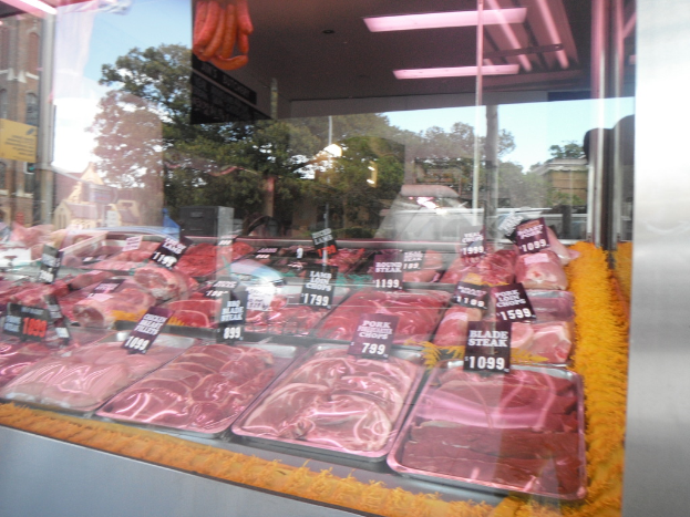 Eine Glasvitrine in einem Metzgerladen, die Tabletts mit Fleisch zeigt, mit Preisschildern, wobei durch das Fenster Gebäude und Bäume sowie die Decke des Raums oben zu sehen sind.