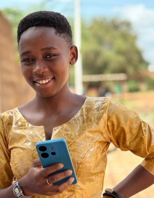 Junges Mädchen in gelbem Kleid mit einem blauen Handy, das es in der Hand hält, und einem Hintergrund aus Wand, Pfahl, Bäumen und Himmel.