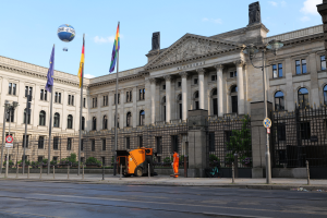 Das Reichstagsgebäude in Berlin, Deutschland, ein großes Gebäude mit zahlreichen Fenstern, umgeben von Straßenlaternen, Lichtern, Schildern, Fahrzeugen, Menschen, Flaggen, Gittern und Bäumen, unter einem bewölkten Himmel.