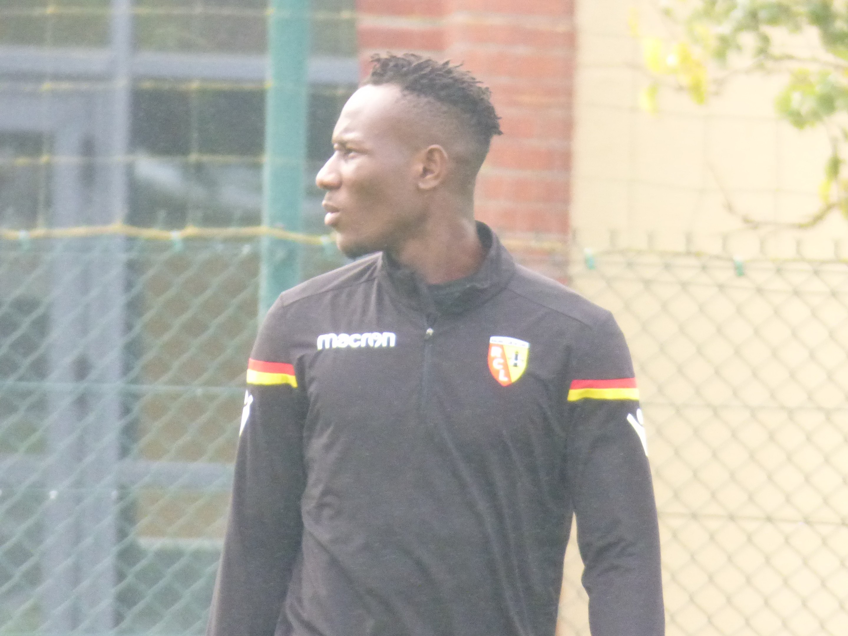 Ein Mann In Einer Schwarzen Jacke Steht Auf Einem Fussballfeld, Umgeben Von Einem Zaun Und Eines Baumes, Und Sieht konzentriert Aus During A Training Session.