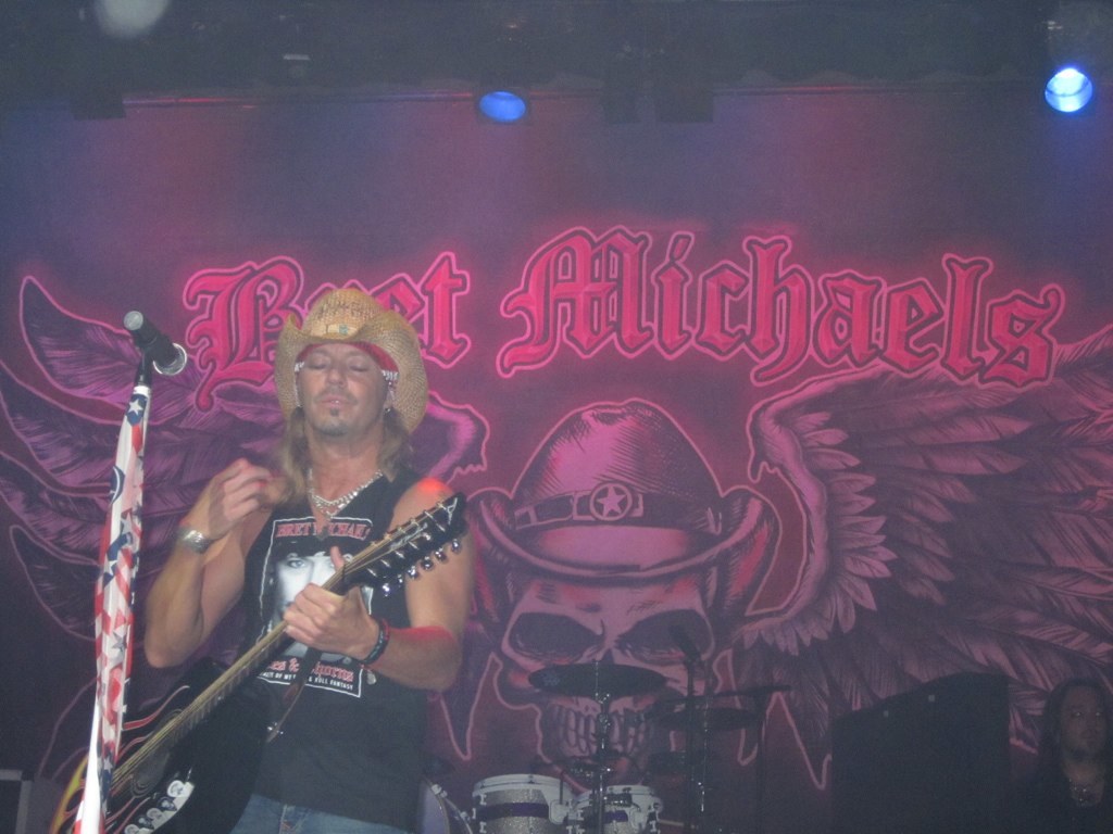 Eine Musik-Konzertszene mit Scheinwerfern oben, einem Banner im Hintergrund, einer Person, die links Gitarre spielt und ein Mikrofon vor sich hat, Drogen in der Mitte unten und einer weiteren Person, die rechts unten sitzt.