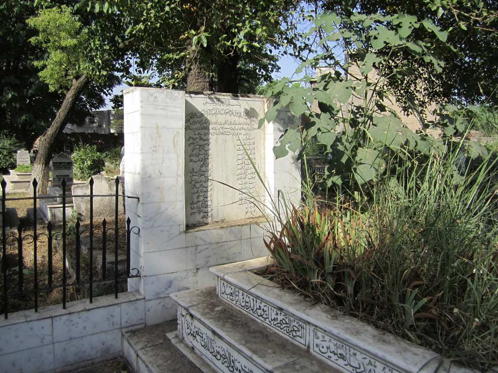 Eine Gedenktafel neben einem Geländer in einem Friedhof, mit Treppen unten, Gras und Bäumen auf der rechten Seite, Gräbern hinter dem Geländer und Gebäuden und Bäumen im Hintergrund.