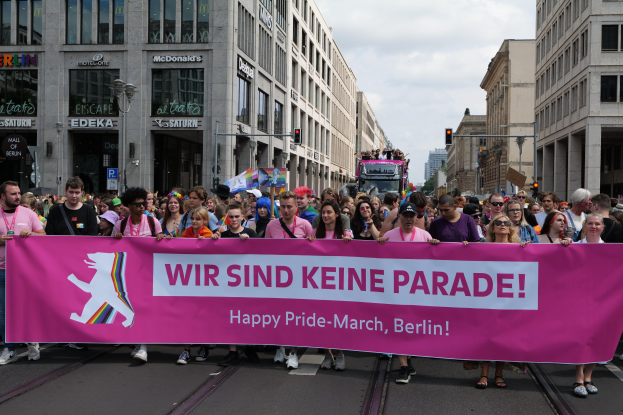 Eine Gruppe von Menschen, die eine pinkfarbene "Happy Pride Parade"-Fahne schwingend durch eine Berliner Straße marschiert, mit Gebäuden, Laternenmasten und Verkehrssignalen an der Straße unter einem bewölkten Himmel.