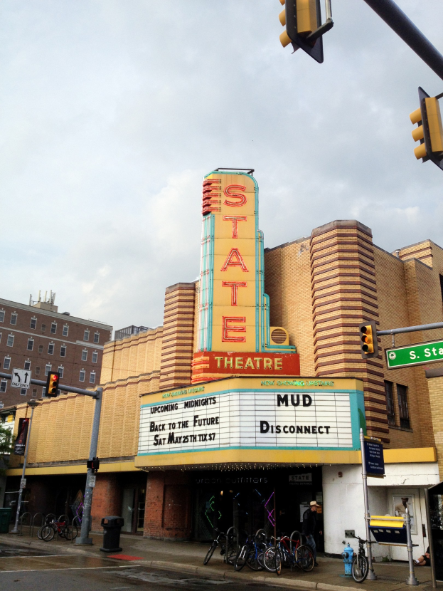 Außenansicht des Staatstheaters in Detroit, Michigan, mit einem Schild, Fahrrädern, einer Person, Pfosten, Ampeln, Schilden, einem Baum und einem bewölkten Himmel.