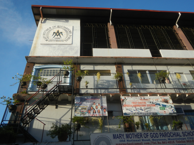 Mariä-Mutter-Gottes-Katholische Schule mit Fenstern, Geländern, Treppen, Topfpflanzen, Bannern und einem bewölkten Himmel im Hintergrund.