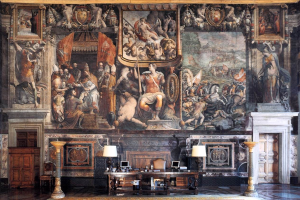 Großer Raum mit einem prominenten Gemälde an der Wand, einem Tisch mit verschiedenen Gegenständen, zwei Lampen zu beiden Seiten des Tisches, zwei Türen auf gegenüberliegenden Seiten und einem gemusterten Teppich auf dem Boden, befindet sich im Palazzo della Signoria in Florenz, Italien.