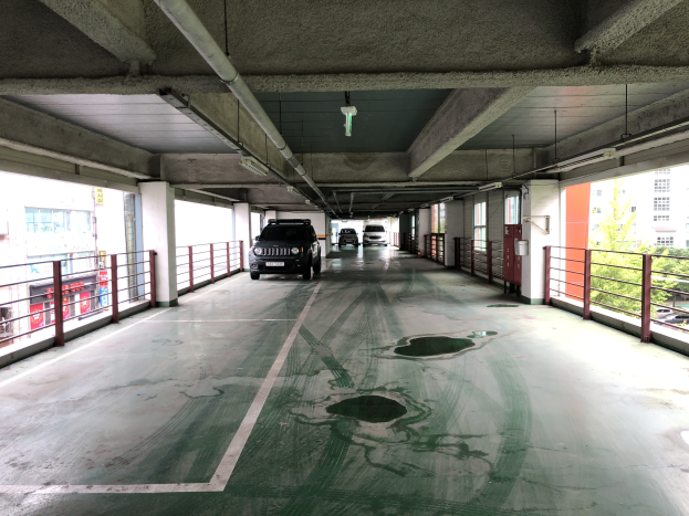 Ein mehrgeschossiges Parkhaus mit parkenden Autos, Geländern, Säulen, Deckenleuchten, freiliegenden Rohren und nahen Bäumen vor einem Hintergrund aus Gebäuden mit Fenstern in einer Stadtlandschaft.