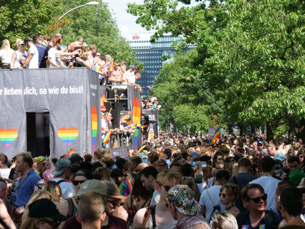 Große Menschenmenge vor Lkw beim Christopher Street Day in Berlin, viele mit Mützen und Schutzbrillen, einige mit Fahnen, Lkw-Banner und Bäume, Gebäude und Laterne im Hintergrund bei bewölktem Himmel.