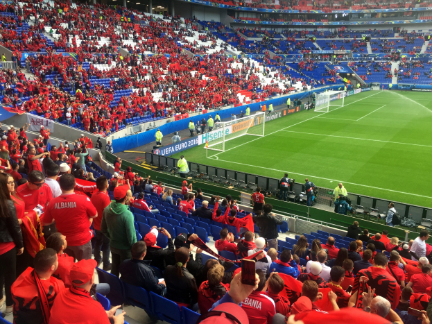 Ein volles Stadion mit Zuschauern, einige sitzen, andere stehen, mit Bannern, einem Torpfosten und Deckenleuchten, während die Menge gespannt auf den Beginn des Fußballspiels wartet.